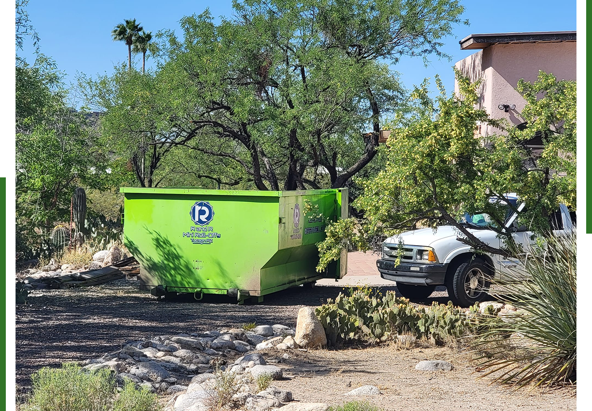 Green dumpster and white truck near trees.